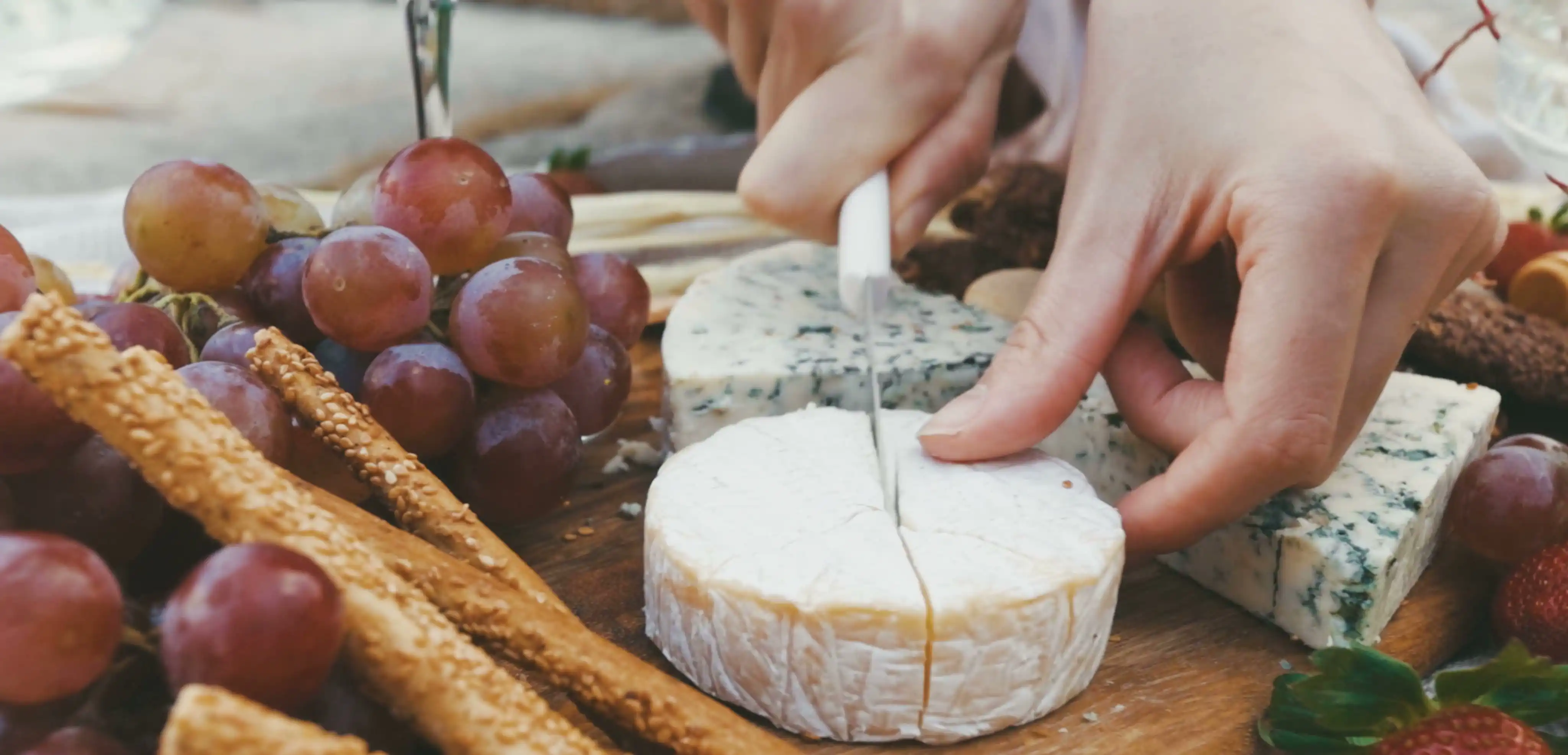 Hands cutting a wedge of cheese beside a bunch of grapes