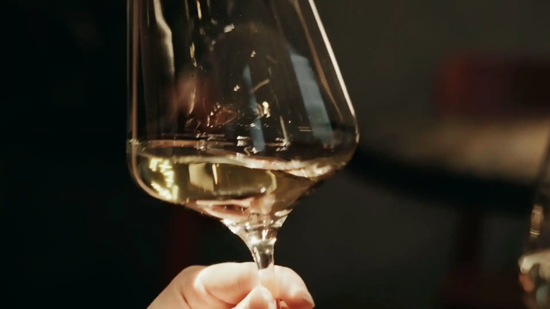 A hand holding a glass of pale white wine against a dark room